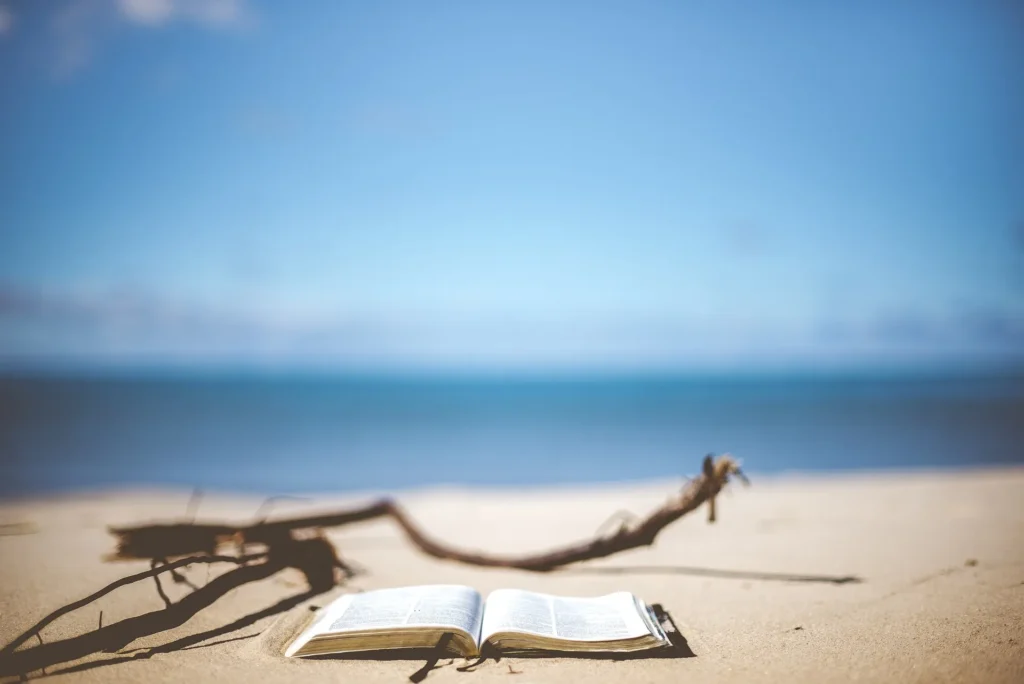 Open book on a sandy beach with the sea and blue sky in the background.